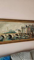 Framed painting showing a bridge, river with boats, and buildings in Paris-style cityscape.