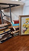 Photo of stacked books arranged on the floor next to framed floral painting under a table, showing titles and variety of books included