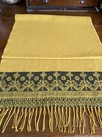 Mustard-colored wool scarf laid flat showing the black decorative motif along edge and fringed end.