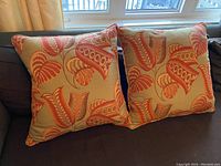 Two feather pillows with 22 inch square covers in pale tea color with tangerine, orange, and muted red flora and fauna motifs, sitting on a sofa near a window.
