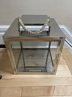 Front angled view of the lantern showing chrome frame, glass panels, hinged door, and thick rope handle on top on wood floor.