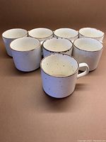 Eight ceramic stackable mugs arranged on brown background, showing exterior with creamy color and dark speckled rims.