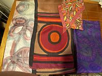 Four silk scarves displayed on a wooden table showing various styles including hand-painted abstract designs and geometric circles.
