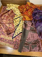 Collection of 5 silk scarves with various prints and colors including purple, yellow, orange, and pink, draped on a wooden surface.