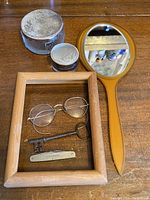 Overview photo showing the collection items arranged on a wooden surface: celluloid hand mirror, two vanity glass jars, round metal framed glasses, wooden frame, old skeleton key, and pocket tool.