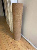 Rolled cork yoga mat standing upright against a wall on a wooden floor, secured with rubber bands.