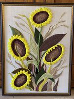 Full view of a framed hand embroidered textile wall art depicting five bright yellow sunflowers with brown centers, green leaves, and beige wheat stalks on cream linen background.
