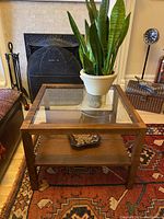 Front angle view of the square coffee/side table showing the solid wood frame, glass top, and teak veneer shelf underneath with a plant and a dish as decor.