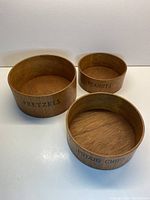 Three teak nesting bowls arranged side by side showing the labeled sides 'PRETZELS,' 'PEANUTS,' and 'POTATO CHIPS.'