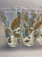 Close-up photo of several clear glass highball tumblers showing detailed gold leaf patterns with turquoise and pale blue leaf elements.