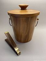 Front view of teak ice bucket with lid and matching ice tongs laid beside it, showing overall shape and size.