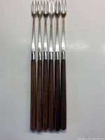 Set of six stainless steel fondue forks laid out in parallel showing full length from prongs to wood handles.