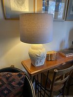 Full view of the textured cream ceramic table lamp with raised woven and flora/fauna motifs, lit with the fabric shade providing warm ambient light in a living space.