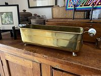 Full view of solid brass trough on wooden surface showing rectangular shape and claw feet.
