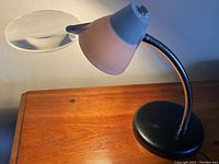Side angle showing the articulating arm, round textured black base, frosty white conical metal shade, and attached round magnifying glass.