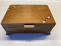 Closed walnut wooden box resting on four feet, with decorative inlay on top corners and visible grain.