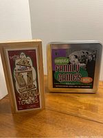 Photo showing the wooden box of Timeless Classic Wood Teasers and the metal tin of Complete Family Games Night side by side.