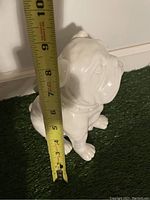 Ceramic white bulldog figurine with tape measure showing about 9 inches tall, placed on carpet against a white wall.