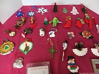Full view of the lot arranged on a red surface showing 30 Christmas ornaments including snowflakes, stockings, trees, musical instruments, characters, plus a handprint ornament craft and some erasers.