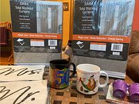 Two packages of SunBlock Total Blackout Curtains with product details on packaging, two coffee mugs, and two reusable purple coffee filters on table.