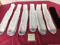 Seven pairs of men's socks laid out on red surface. Six pairs are white with gray toe and one black pair on far right. Socks have Hanes logo on toe area. Photo shows full length of socks.