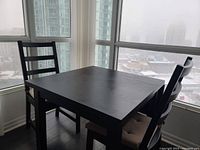 View of black expandable IKEA dining table and two chairs with beige cushions in a corner by large windows in a high-rise apartment.