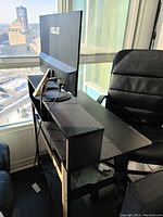 Side view of black multi-level office table showing the top level with an ASUS monitor on it, middle workspace surface, and lower shelf storage area. Chair and window background visible.