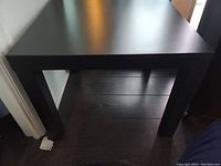 Side view of the black square side table showing one corner with leg and part of floor underneath, highlighting the table's sturdy construction and matte black finish.