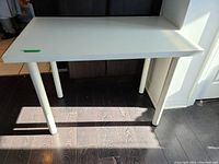 White study table shown from a side angle with four cylindrical white legs, placed on dark wood flooring, some sunlight casting shadows.
