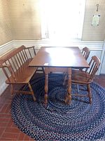 Full view of the vintage wooden dinette set including the drop leaf table, bench, and two chairs positioned on a braided rug under a window with yellow checked wallpaper.