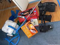 Photo showing leg brace, blood pressure monitor box, heating pad with blue tubing, wireless noise reduction headset, polar care cooling wrap, and other items on floor