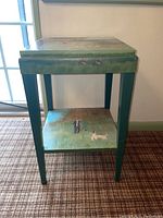 Full view of painted side table showing square shape, greenish teal legs, top surface artwork and lower shelf painted scene.