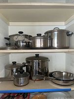 Shelving with six Revere Ware stainless steel pots with lids and two pans, black handles and knobs, various sizes