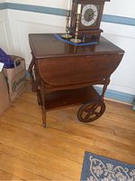 Side view of vintage wooden tea cart with drop leaf top, paired with two large rear wheels and a lower shelf, placed on hardwood floor.