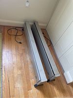 Two rectangular metal space heaters with handles and power cords laying on wooden floor, shown from top angle.