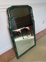 Full view of the mirror showing green ornate frame with hand-painted scenes and floral motifs, leaning against white wall and reflecting furniture.
