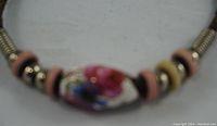 Close-up of white porcelain bead with pink floral motif and surrounding metal and disk beads on leather band bracelet.