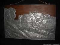 Large hand hammered pewter metal on wood wall hanging depicting houses by water, signed J Hebert, measuring 21 by 14 inches.