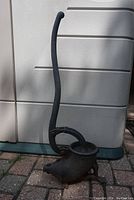 Full view of the vintage cast iron hand pump showing the curved handle, spout, and base on a brick surface near a storage container.