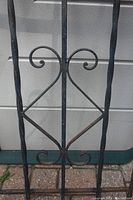 Close-up of wrought iron fence section showing decorative twisted vertical bars and symmetrical scrolls in the center diamond shape.