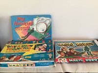 Three vintage board games: My Hankies Embroidery Set, Give-a-Show Projector, and The Amazing Magic Robot, arranged on a white surface.