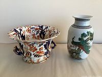 Pair of oriental style ceramic pieces, floral pedestal pot with handles and peacock vase side by side on neutral background.