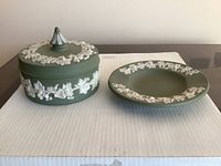 Photo of the green Wedgwood tobacco jar and matching ashtray, showing white jasperware relief floral and grapevine decoration on matte green background.