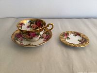 Set showing tea cup on saucer and separate ashtray with golden rose pattern and cracked saucer.