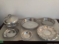 Top view of all six silverplated servingware pieces arranged in a row on a white surface