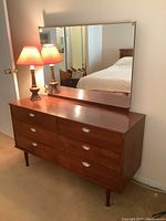 Full front view of wooden dresser with mirror, lamp on left, and reflection showing bed in background.