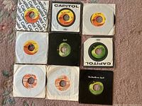 Top view of all nine 45 RPM vinyl record singles with original sleeves, showing label and sleeve variety including Capitol, Apple, and London.