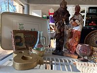 Photo showing two tall figurine sculptures of people in traditional clothing, a white metal serving tray with leaf design, a vintage tin container, a glass pitcher with floral design, and a gold wooden ampersand symbol.