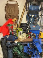 Overall assortment of camping gear including backpacks, camping chairs, lanterns, sleeping bags, and various accessories arranged on and around tables and containers.