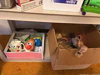 Photo of box with various children's toys and mugs including a Mr. Potato Head toy and Minnie Mouse mug under a beige plastic shelf
